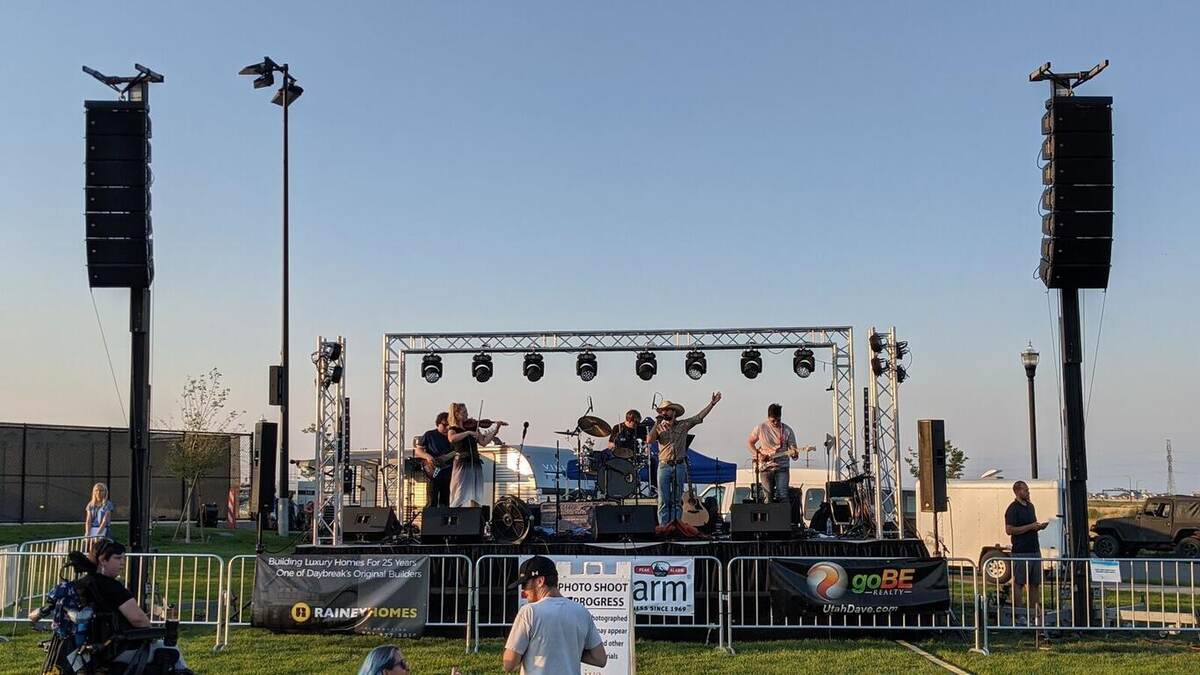Daytime outdoor community-event stage with truss goalposts, stacked line arrays and subs, a band performing, and safety-fenced crowd area under a clear sky