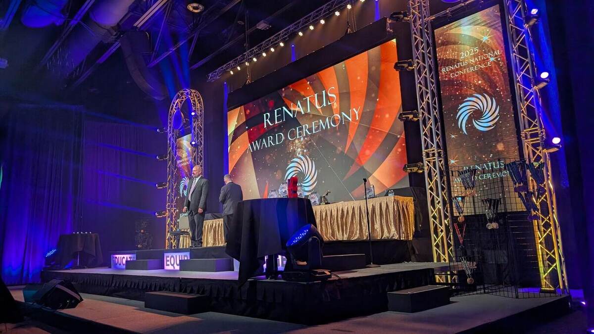 A 'Renatus Award Ceremony' stage with a large curved LED screen, black truss framing, a head table, warm/blue lighting, and two speakers standing at stage left