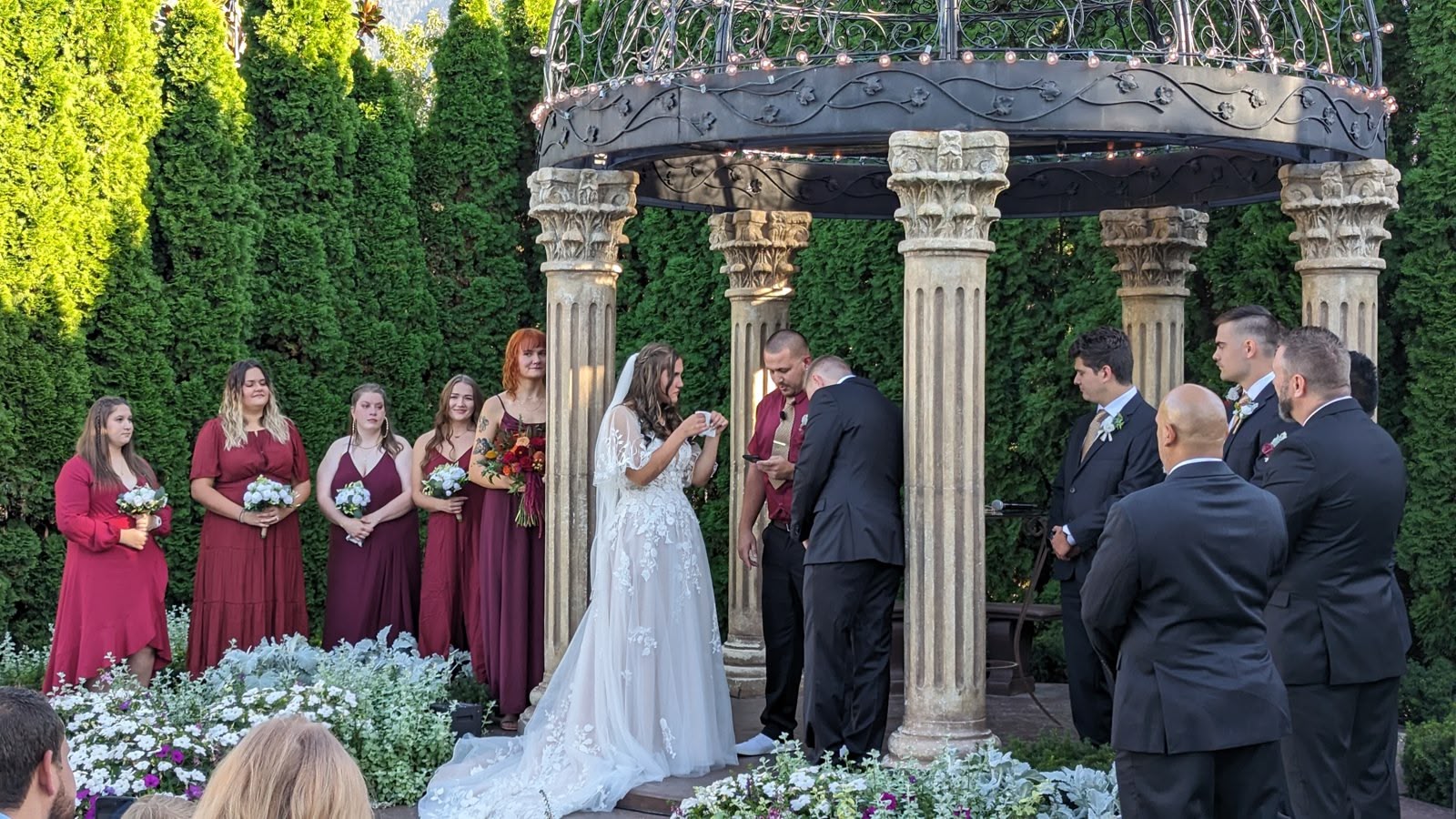 An outdoor garden wedding ceremony under an ornate columned gazebo, the couple exchanging rings as the wedding party looks on